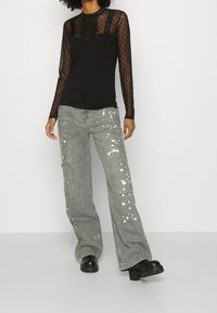 Schwarzes Langarmoberteil mit transparenten, gepunkteten Mesh-Ärmeln, kombiniert mit grauen, weit geschnittenen Jeans mit weißem Farbspritzer und Cargo-Taschen.
