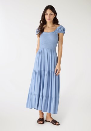 Donna che indossa un vestito midi azzurro a balze con maniche corte a sbuffo e scollo quadrato, abbinato a sandali aperti marrone scuro.