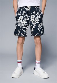 Marineblå shorts med hvitt blomstermønster, med elastisk liv, sidelommer og avslappet passform. Hvite joggesko med marineblå detaljer og stripete sokker.