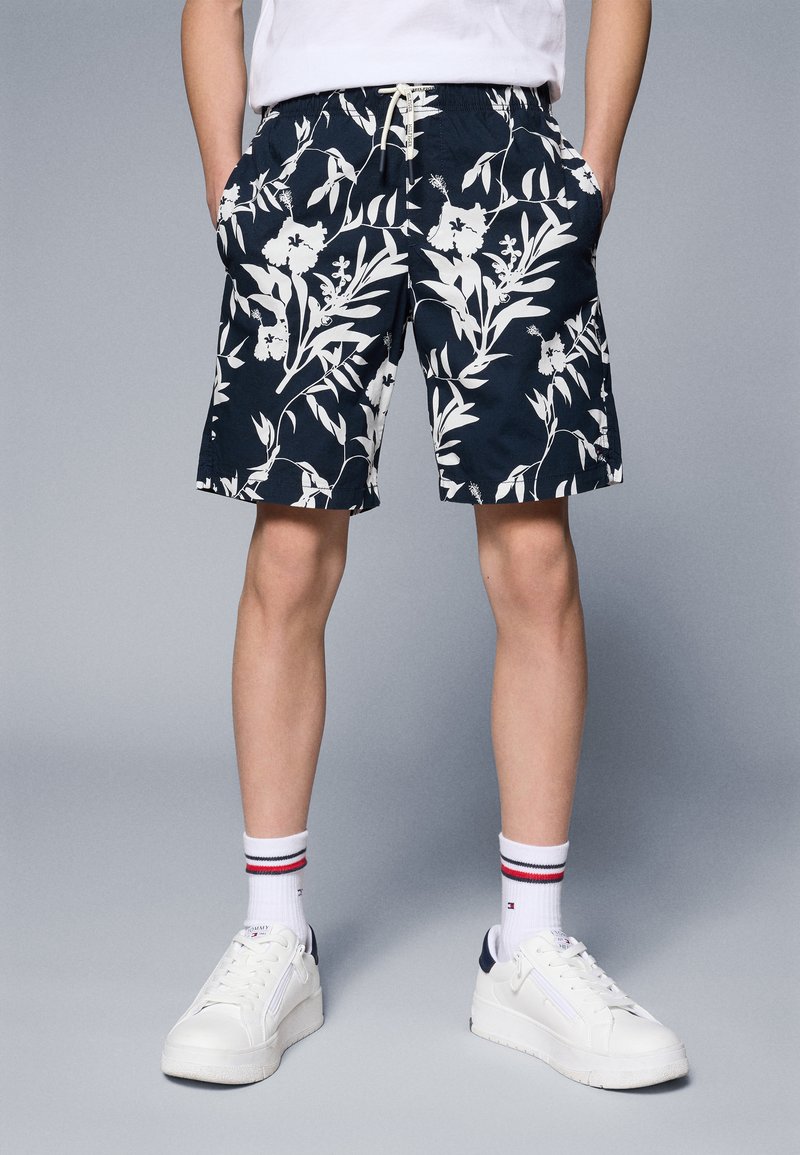 Marineblå shorts med hvitt blomstermønster, med elastisk liv, sidelommer og avslappet passform. Hvite joggesko med marineblå detaljer og stripete sokker.