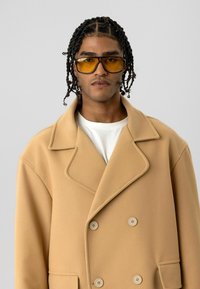 Beige double-breasted jas met grote revers, knopen en voorzak. Gedragen over een wit overhemd, gecombineerd met geel getinte zonnebril.