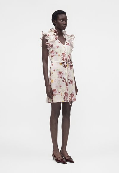 Γυναίκα με αμάνικο φόρεμα με λουλούδια, βολάν στους ώμους και ζώνη για δέσιμο, σε συνδυασμό με σκούρα κόκκινα μυτερά παπούτσια με τακούνι, στέκεται μπροστά σε λευκό φόντο.