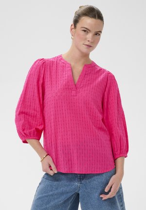 Femme portant un chemisier texturé rose vif avec des manches bouffantes trois-quarts et un jean bleu, debout avec une main dans la poche devant un fond uni.