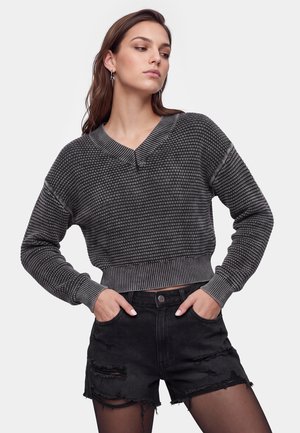 Kvinde iført en sort og grå strikket sweater og sorte revnede denimshorts med hænderne i lommerne, kigger til siden mod en ensfarvet baggrund.