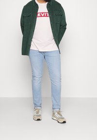 Chemise en velours côtelé verte, t-shirt graphique rose, jean bleu clair et baskets beige avec des accents gris et blancs. Debout devant un fond blanc.