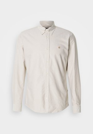 Világos bézs színű, hosszú ujjú, gombos ing hegyes gallérral és egy kis hímzett logóval. Puha anyag, sima textúrával és egyenes szegéllyel.