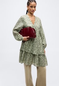 Γυναίκα με πλεξούδες, ντυμένη με πράσινο φόρεμα με φλοράλ μοτίβα και μπεζ παντελόνι, κρατώντας μια μικρή βουργουνδί δερμάτινη τσάντα σε απλό φόντο.