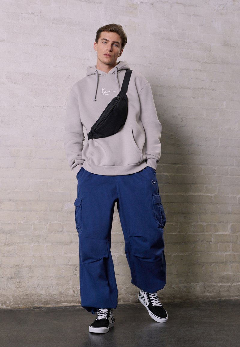 Haut en molleton gris clair avec cordon de serrage, pantalon cargo bleu marine avec poches, sac banane noir, et chaussures à enfiler noir et blanc à carreaux.