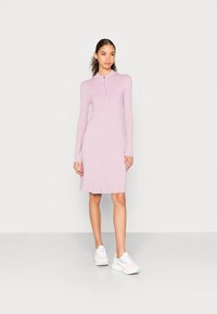 Vestido rosa claro com nervuras, colarinho com fecho de meia-zíper, mangas longas, bainha rodada e textura subtil. Combinado com sapatilhas brancas.