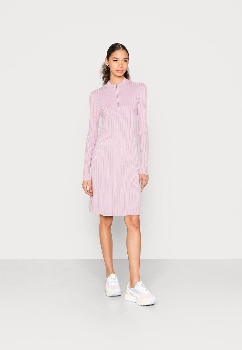 Vestido rosa claro com nervuras, colarinho com fecho de meia-zíper, mangas longas, bainha rodada e textura subtil. Combinado com sapatilhas brancas.