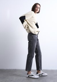 Γυναίκα που στέκεται φορώντας καφέ πουλόβερ με μαύρα επιθέματα στους αγκώνες, γκρι ψηλοκάβαλα τζιν και λευκά αθλητικά παπούτσια σε τσιμεντένιο πάτωμα μπροστά σε άσπρο τοίχο.