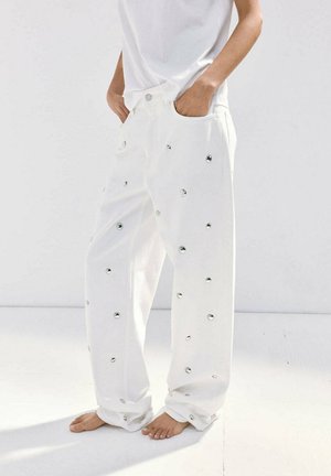 Persona con pantalones blancos holgados con tachuelas circulares reflectantes, descalza y una camisa blanca sencilla, de pie frente a un fondo blanco.