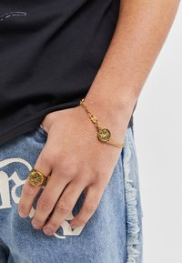 Gold jewelry set includes a bracelet with a circular pendant and a chunky ring, both featuring intricate designs. Worn on a hand against denim.