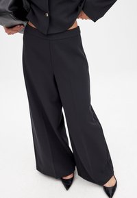 Melnas platās bikses, izgatavotas no gludas auduma, ar augstu vidukli un sānu rāvējslēdzēju. Apvienotas ar norādītiem melniem zābakiem.