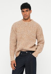 Pulover din tricotaj gros, de culoare maro deschis cu stropi albi, cu gât rotund și mâneci lungi, asortat cu blugi negri din denim.