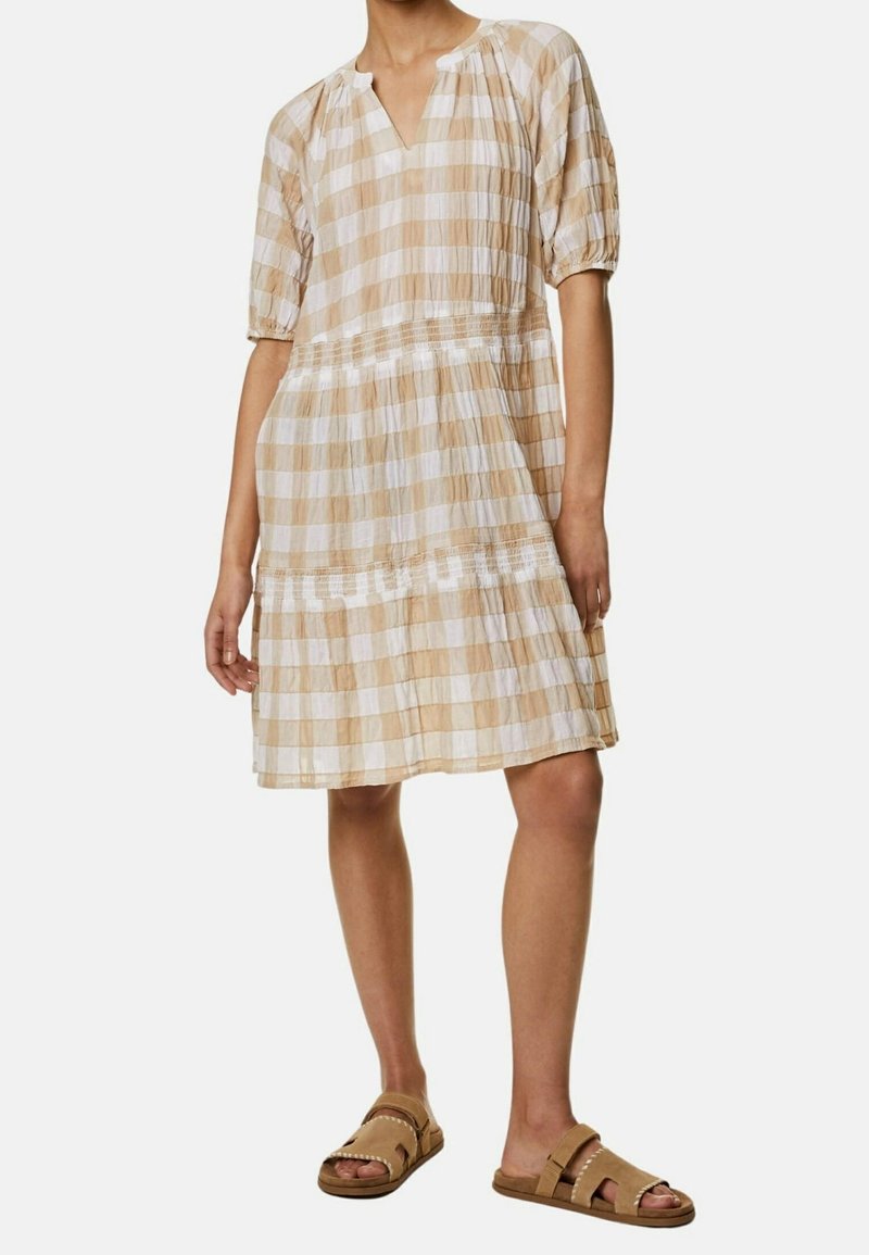 Femme portant une robe à carreaux beige et blanche, longueur genou, avec des manches bouffantes et des sandales ouvertes couleur marron, debout devant un fond blanc.