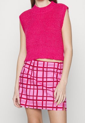 Frau trägt ein ärmelloses, knallpinkes gestricktes Croptop und einen pink karierten Minirock mit roten Linien und kleinen vorderen Schlitzen.