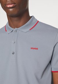 Polo gris avec des accents rouges au niveau du col et des manches. Il possède trois boutons et un logo "HUGO" brodé en rouge sur la poitrine gauche.