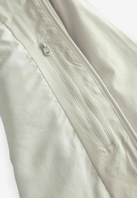 Light beige jacket fabric with a smooth texture, featuring a vertical zip and a fabric overlay with subtle sheen.