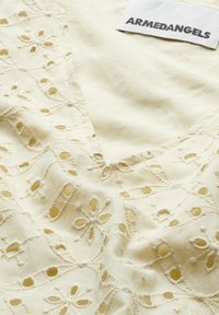 Weißer Baumwollstoff mit floralem Stickerei und Ausschnitten. Verfügt über einen V-Ausschnitt und ein Etikett mit der Aufschrift "ARMEDANGELS". Weiche Textur.