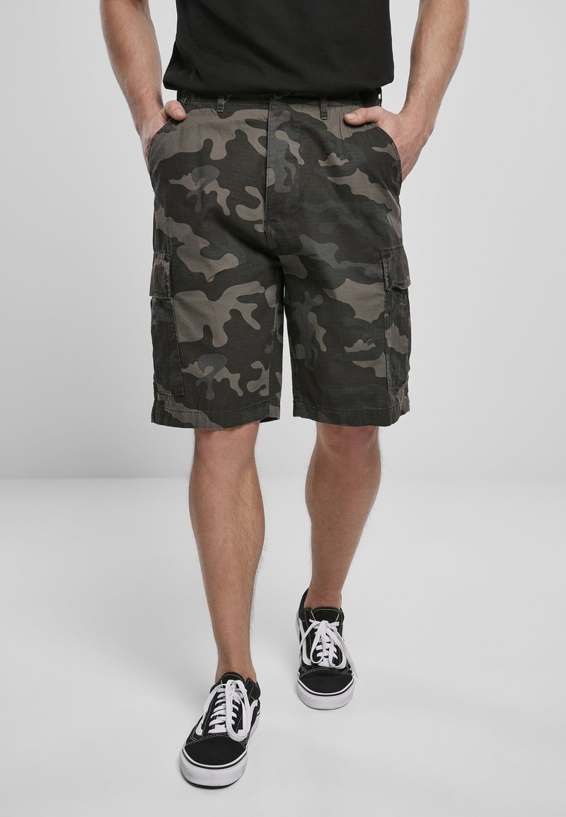 Homme portant un short cargo camouflage noir et gris avec des poches, un t-shirt noir et des baskets noires avec des lacets blancs, debout les mains dans les poches.
