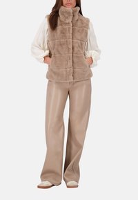 Chaleco de piel sintética en suave tono beige con un collar alto, diseño acolchado y bolsillos delanteros. Combinado con pantalones de pierna ancha beige lisos y calzado ligero.