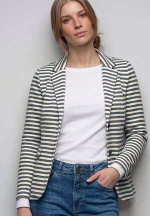 Mujer con cabello castaño claro recogido en un moño bajo, vistiendo una camisa blanca, vaqueros azules y un blazer azul marino con rayas blancas, de pie frente a un fondo gris.