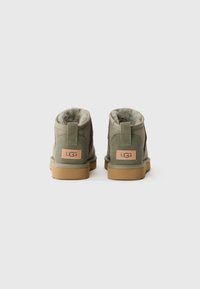 Par de botas UGG de ante gris con suelas color beige y forro de piel de oveja, vistas desde atrás sobre un fondo blanco.