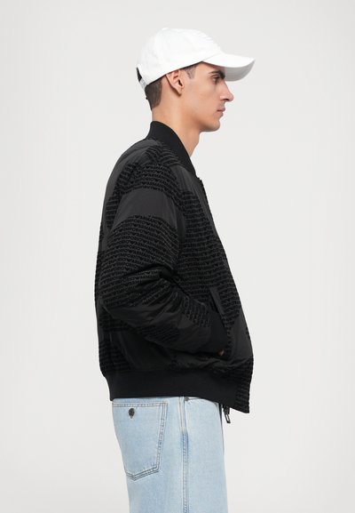 Black bomber jacket with a textured pattern featuring the brand name, ribbed cuffs, and a relaxed fit, paired with light blue jeans.