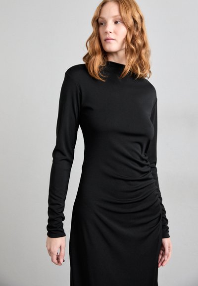 Robe longue noire à manches longues en tissu lisse, présentant un col haut et des détails froncés sur les côtés pour une texture supplémentaire.