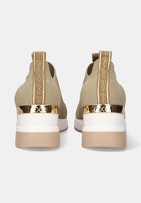 Zapatillas de punto beige con suelas de goma grises, acento dorado en el talón y detalle del logo en la pestaña de tirón. Presentan un diseño elegante y redondeado.