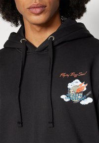 Fekete kapucnis felső, amely húzózsinórral rendelkezik, élénk grafikai ábrázolással szárnyakról és felhőkről. A szöveg: "Repülő Bzzz Saw!" és "Alpha Industries."