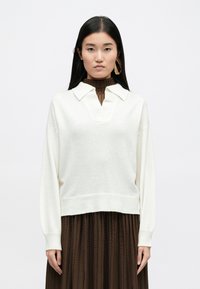 Λευκό πλεκτό πουλόβερ με V λαιμόκοψη, ραβδωτό γιακά και μακριά μανίκια, συνδυασμένο με μια πτυχωτή καφέ φούστα, αναδεικνύοντας υφές υφασμάτων.