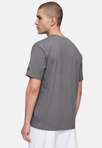 Camiseta gris de mangas cortas, de material de algodón, diseño de cuello redondo, textura suave, lisa sin patrones, con un logo en la manga izquierda.
