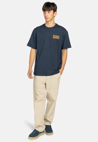 Navyblauwe T-shirt met een geel logo op de borst, gecombineerd met lichtbeige broek en blauwe schoenen. Eenvoudig ontwerp, korte mouwen, relaxte pasvorm.