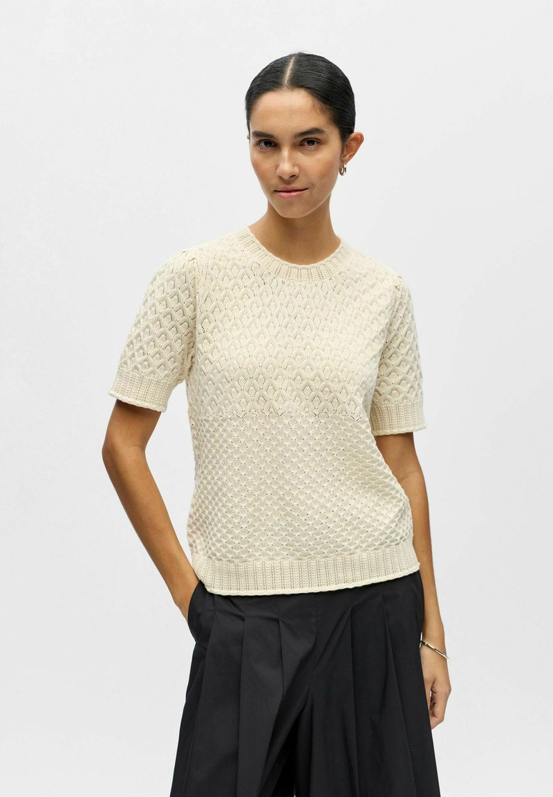 Femme aux cheveux foncés portant un haut en maille crème à manches courtes et un pantalon plissé noir, debout avec une main dans la poche sur un fond uni.