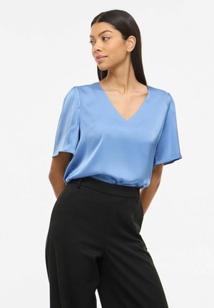 Femme aux longs cheveux noirs portant un chemisier bleu clair en satin à col en V et un pantalon noir taille haute, regardant vers sa gauche.