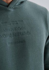 Kapuzensweatshirt in Dunkelgrün, mit geprägtem Text: "AVIATOR ADVENTURE", "TESTED AT HIGH ALTITUDE" und "PME LEGEND." Aus weichem Material gefertigt.