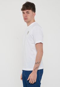 T-shirt bianco in cotone con maniche corte, colletto rotondo e una piccola etichetta con logo sul lato sinistro del petto. Indossato con jeans blu.