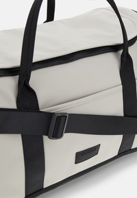 Borsa a viaggio impermeabile bianca con dettagli neri, dotata di chiusura a cerniera, manici piatti e una tasca esterna con patch del logo.