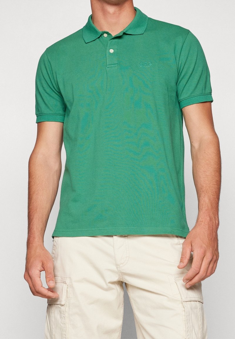 Homme portant un polo vert à manches courtes avec le logo "GAP", associé à un pantalon cargo beige, debout devant un fond uni.