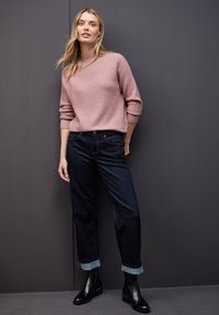 Kobieta w różowym swetrze o prążkowanej strukturze, ciemnych dżinsach z podwiniętymi nogawkami i czarnych botkach za kostkę, stojąca przy ciemnoszarej ścianie.