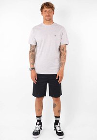 T-shirt gris à manches courtes avec un logo minimaliste sur la poitrine gauche, associé à un short noir et des baskets montantes noires. Peau visible sur les bras.