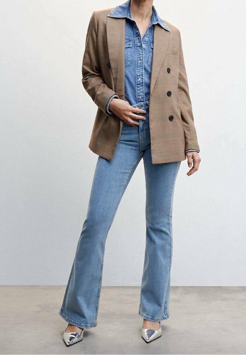 Personne portant une chemise en denim bleu clair et un jean avec un blazer croisé à carreaux marron et des talons pointus argentés, debout sur un sol en béton.