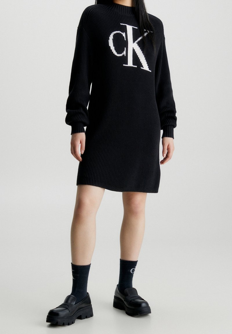 Femme portant une robe-pull côtelée noire avec le logo blanc "CK", des chaussettes noires avec des inscriptions blanches et des chaussures à plateforme noires épaisses.