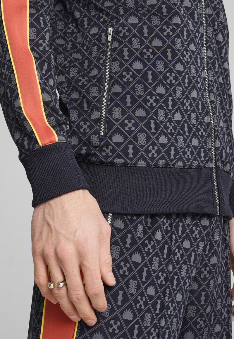 Giacca a zip blu navy e pantaloni coordinati presentano un motivo geometrico con accenti grigi. Strisce rosse e gialle sulle maniche, polsini a coste testurizzati.