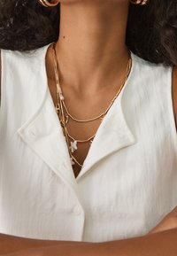 Biała bluzka zapinana na guziki o teksturowanym wykończeniu, nałożona z wieloma złotymi naszyjnikami o różnych długościach i dekoracyjnymi zawieszkami.
