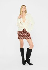 Camicia crema abbottonata con una profonda scollatura a V, legata in vita, abbinata a una gonna corta a scacchi rosso e bianco e stivali neri con tacco al ginocchio.