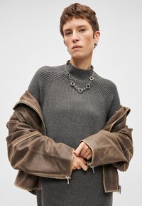 Maglione grigio lavorato a maglia con colletto alto, indossato sotto una giacca in pelle marrone oversize con polsini con zip e una texture liscia. Collana in catena metallica.