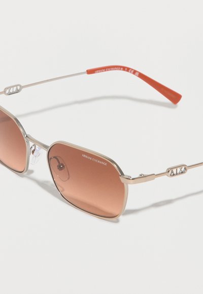 Lunettes de soleil avec monture en métal doré, verres dégradés marron et embouts de branches rouges, arborant la marque Armani Exchange sur les verres et les branches.